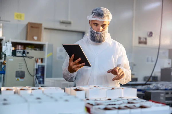 白い滅菌ユニフォームの監督者が食品工場に立っていて 収集されたクッキーで箱を数えています 彼はタブレットを持っていて労働者の生産性をチェックしている — ストック写真