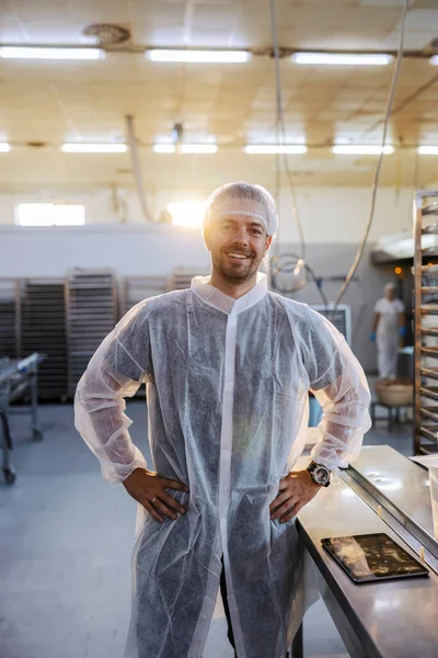 A successful food factory owner in a white sterile uniform is standing in his factory with hand on hips and looking at the camera. He is very happy with the quality of the products.