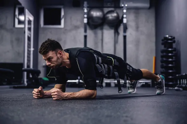 Full body burning exercise, using new EMS technology. A young attractive man in clothes for EMS is in a plank position on elbows in the gym. Electrical muscle stimulation