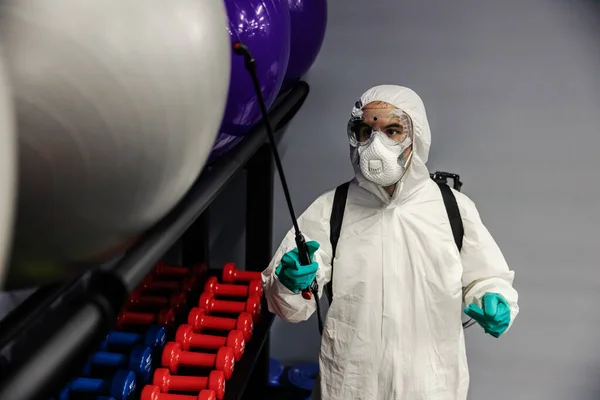 Suppression of COVID-19 virus and cleaning of exercise equipment in the gym by a male person in a protective white suit. The focus of the photo is on the pilates and yoga balls
