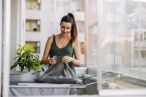 一个年轻女人在阳台上做家务活 堆放衣服和洗衣服的画像 一个穿着休闲装的微笑的女人在阳光灿烂的夏日把毛巾放在阳台上 — 图库照片