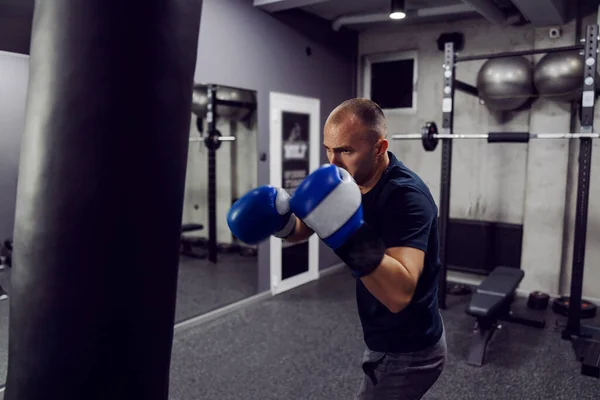A strong hit in the punching bag. A focused man wears boxing gloves and sportswear in an indoor modern gym with equipment. A man who does boxing is just about to hit a huge black punching bag