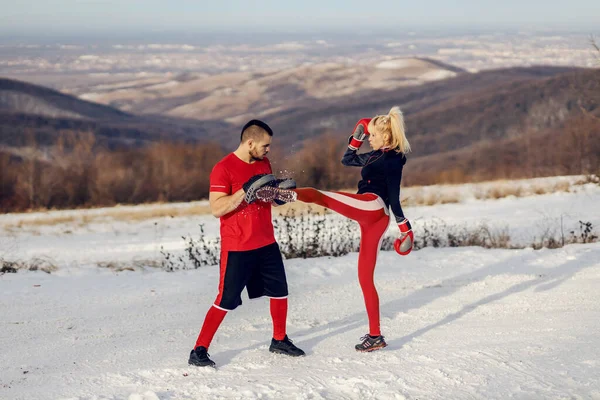 Sportswoman sparring with boxing gloves in nature at snowy winter day with her instructor. She is kicking pad. Boxing, winter fitness, outdoor fitness