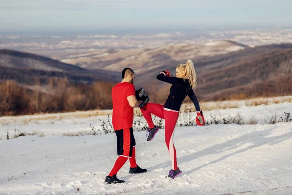 Sportswoman sparring and kicking pad in nature at snowy winter day with her coach. Boxing, winter fitness, outdoor fitness