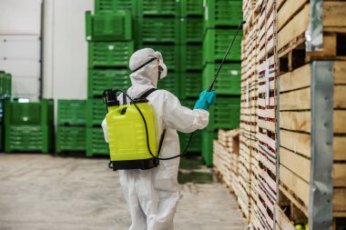 Stay healthy, warning for COVID 19, cruel fight with the coronavirus. A closeup of a person in protective uniform with sprayer prevents the spread of the infection in high-risk spot in warehouse clipart