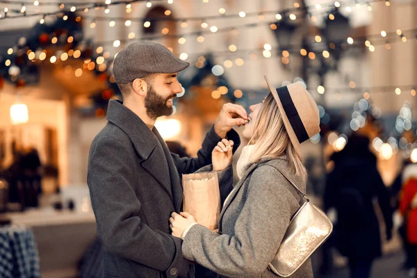 ロマンチックなクリスマスカップルは クリスマスの日に通りにポップコーンを食べます クリスマスの通りに立っている間に若い幸せな男はポップコーンで彼の愛するガールフレンドを養う — ストック写真