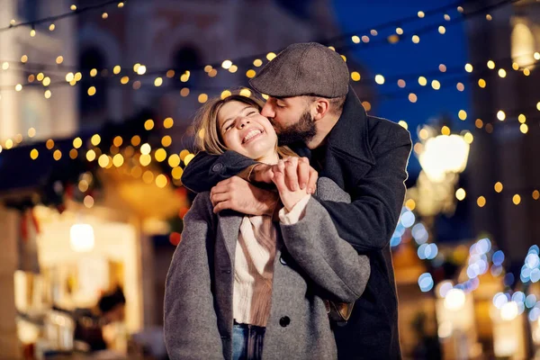 クリスマスカップルは新年に通りでキスをします クリスマスの前夜に路上で彼女の最愛のガールフレンドと一緒に立って恋に若い男 彼女を抱きかかえて頬にキス — ストック写真