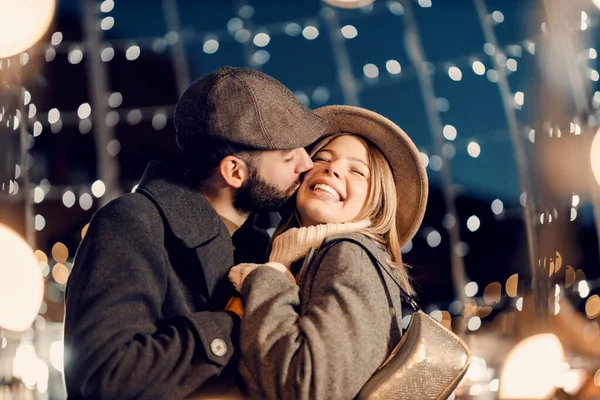 クリスマス愛好家は大晦日に屋外でキスをします 光に囲まれたクリスマスの前夜に屋外に立っている間 若い男は彼の最愛のガールフレンドを抱き 頬にキス — ストック写真