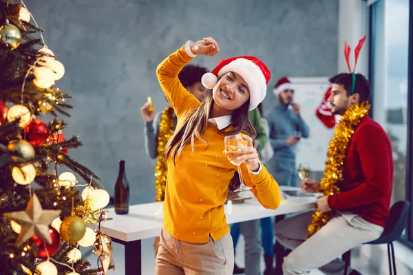 新年のお祝い クリスマスツリーの隣のオフィスで踊っていると 陽気なビジネスマンの女性がシャンパンを持っています ぼやけた背景で彼女の同僚は楽しい時間を過ごしています — ストック写真