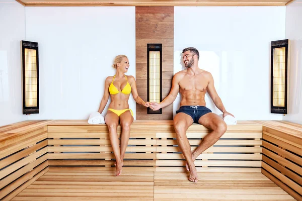 A happy attractive couple in swimwear is sitting in a sauna, holding hands and flirting. Lovely couple spending their anniversary at spa center. A couple holding hands in the sauna