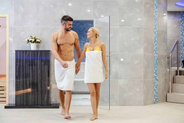 A happily married couple in towels is holding hands and walking in the spa center. They spending quality time together at the resort. Wellness and healthy life concept. Couple at spa center