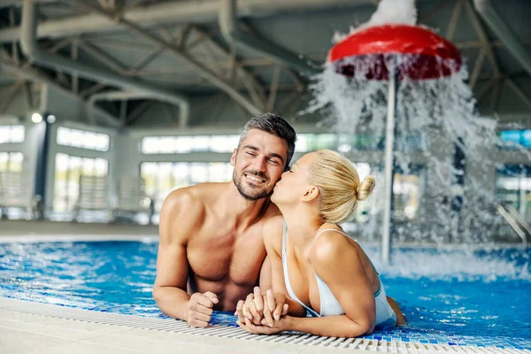 水着姿の金髪女性が温泉センターの温水とミネラルウォーターでプールに立っている間 頬に彼女のパートナーにキスをします カップル接吻でザ プール — ストック写真