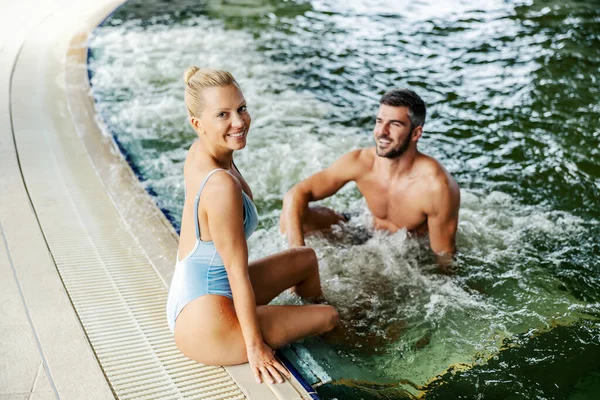 温水プールの隣にセクシーなブロンドが座ってカメラを見ている 彼女のボーイフレンドはプールに座って笑顔 スパセンターのプールで幸せなカップル — ストック写真