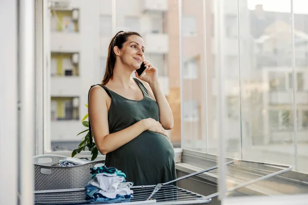 妊娠中の主婦が電話を使って家事をしながら話し笑います 祝福された期待の母親は カジュアルな服でテラスに立って 電話を使用します — ストック写真