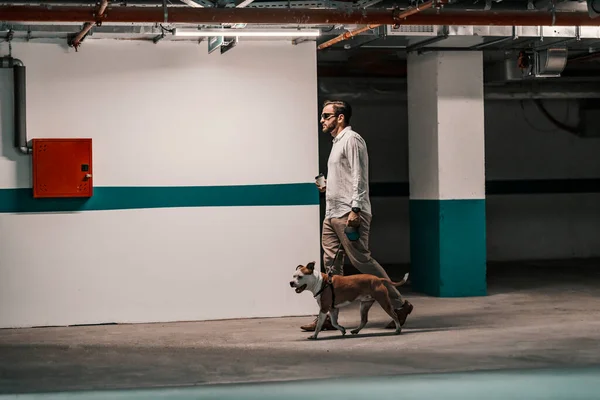A busy man dressed smart casual is searching for his car in the underground garage with his dog. The man is holding coffee to go and passing by the wall.
