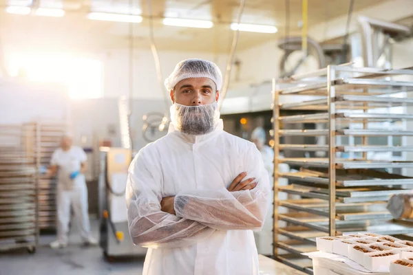 白い滅菌ユニフォームの成功した食品工場の監督者が腕を組んで工場に立っています 彼は製品の品質と量に非常に満足しています — ストック写真