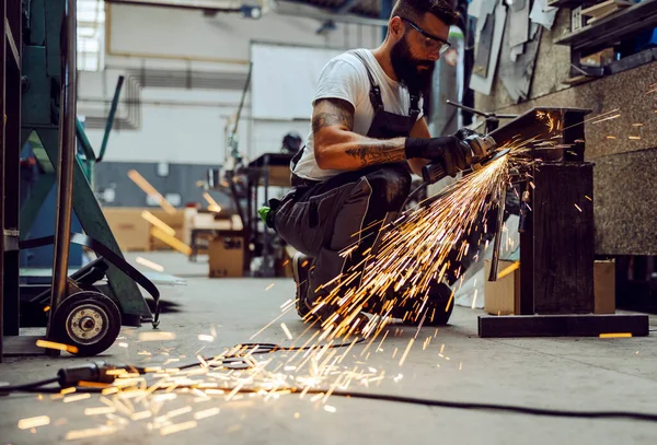 A metal worker kneeling next to metal parts and processing them with a grinder. Worker grinding in the workshop