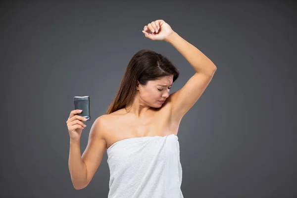 Unpleasant armpit odor. An adult female wrapped in a white towel around her body stands in front of a gray wall and smells the stink of her armpits before applying a deodorant stick. Care and hygiene