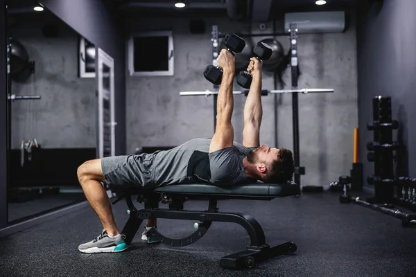 Man keeping good physical shape and fitness. Lifting dumbbells with both hands indoor gym with gray interior. Sports routine and healthy lifestyle. The concept of healthy living, fitness goal