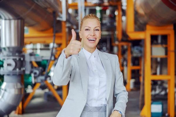 Smiling successful independent female CEO in an elegant and modern suit standing in heating plant and showing thumbs up and looking at the camera. The woman giggles and shows a sign of approval