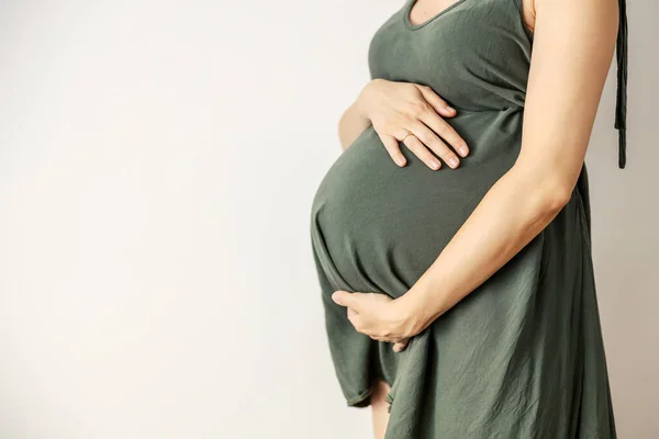 The pregnant woman\'s belly is in focus. The big belly of the future mother is in front of the white background. A woman in a casual gray-green dress stands and holds to her stomach. Motherhood