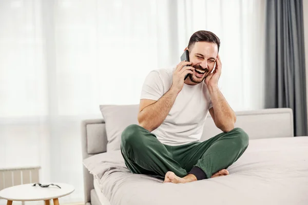一个快乐的人在舒适的家里坐在床上 一边笑一边打电话 — 图库照片