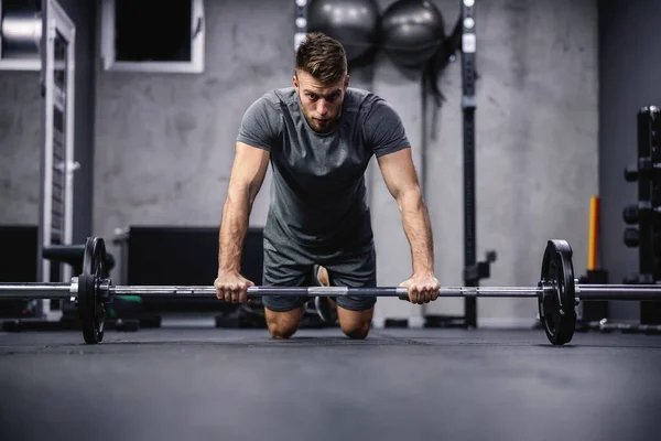 Focus on training and concentration on exercise. A young attractive man in a gray T-shirt rests his hands on a barbell and squats in the gym. Initial body posture and sports discipline