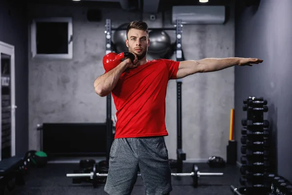 Proper posture of the body and arms when exercising, sports discipline. Attractive man raises a kettle bell over his head in the gym. He holds weight in one hand while he stretches out his other hand