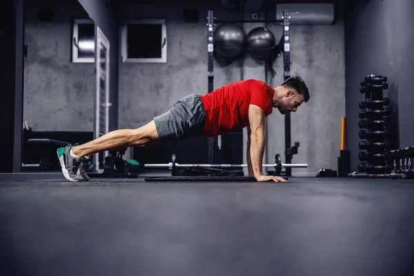 Exercises for arms and shoulders. A man in sportswear with muscles in a modern gym concept does push-ups. He is in a plank position. Empty space for an advertising message