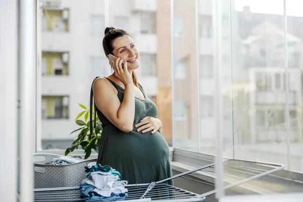 家事をしながら携帯電話を話す妊婦 赤ちゃんの服がワイヤーに乗っている間 妊婦さんはカジュアルな服でテラスに立って電話を使用しています — ストック写真