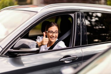 A happy nerdy girl is driving a car and showing thumbs up to other drivers. It is not hard to be polite in traffic. A girl driving a car clipart