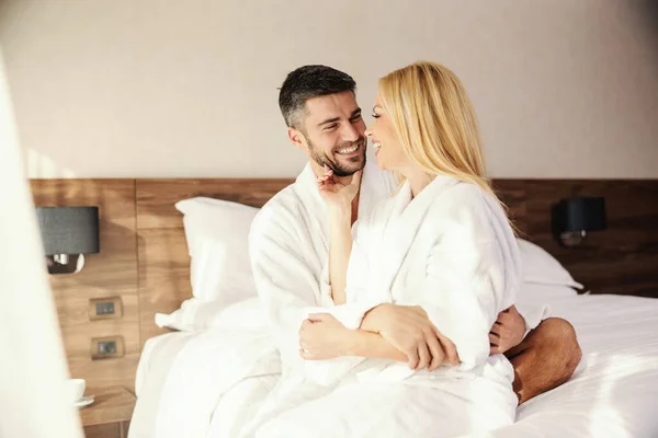Honeymoon, freshly married couple kissing and tenderness in the warmth of a comfortable bed with white sheets. She sits on his lap and he hugs her. Giggles and talks in hotel room, gentle touch
