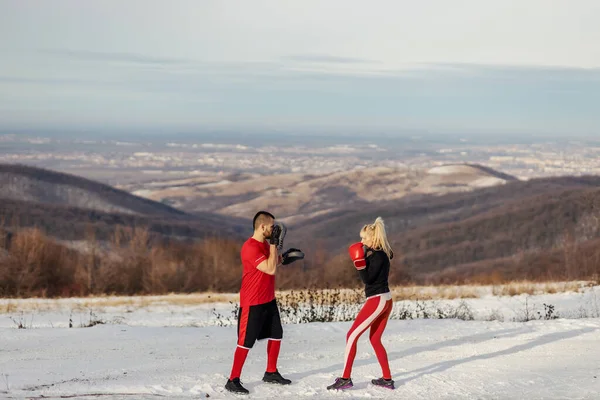 Sportswoman sparring with boxing gloves in nature at snowy winter day with her coach. Boxing, winter fitness, outdoor fitness