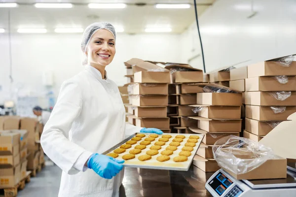 幸せな女性食品工場労働者ベーキングビスケットと笑みを浮かべてトレイを保持カメラ — ストック写真