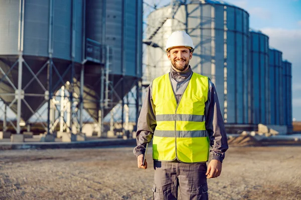 Portrait of industry worker holding tablet in front of silos full of grain and smiling at the camera.