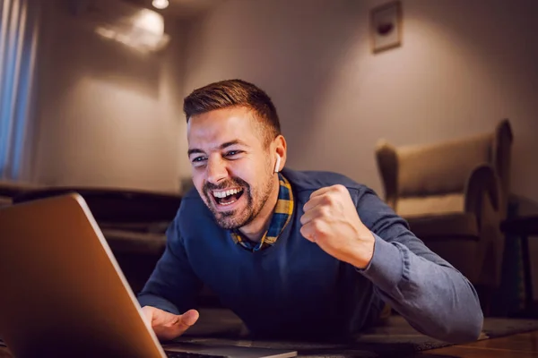 A freelancer celebrates accomplishment and laughing at the laptop while lying down on the floor at home.