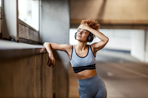 A tired, fit urban runner with headphones standing and wiping the sweat from the forehead while listening to music and taking a break. An urban runner resting.