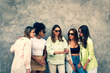 Duvara dayanmış, güneş gözlüğü takmış bir grup gülümseyen genç bayan arkadaş - açık havada genç kadınların portresi - beş genç bayram giysisi ve cep telefonu görünümlü teknoloji kavramı