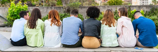 Multi ethnic young people sitting on a bench in a park from behind - shot from behind their backs - Wait concept  group person from back side  look from behind  rear view -  view of people behind pub - horizontal banner or header
