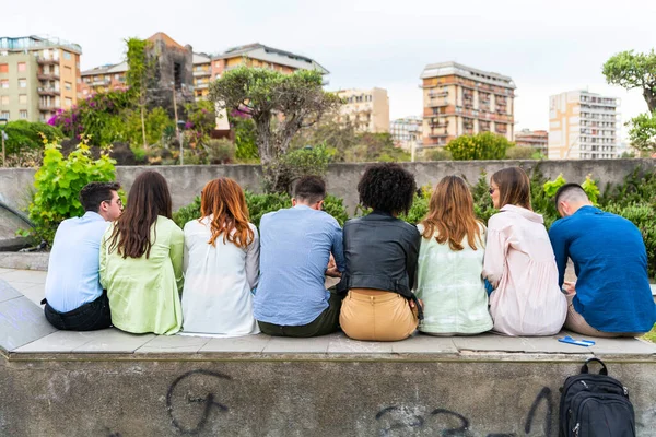 Multi ethnic young people sitting on a bench in a park from behind - shot from behind their backs - Wait concept  group person from back side  look from behind  rear view -  view of people behind  friends from behind