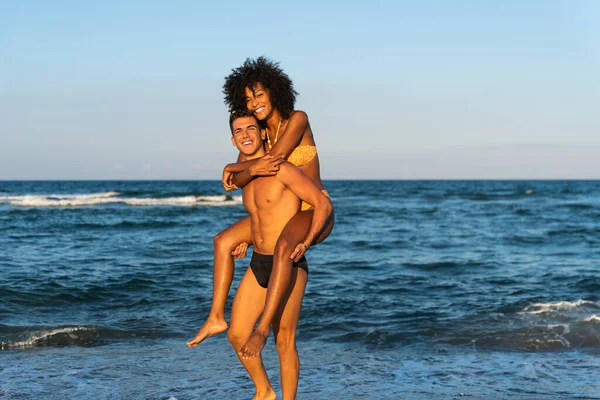 Happy young interracial couple in piggybak on beach  man giving piggybak his woman and laughing at tropical beach  smiling boyfriend in love carrying on back her girlfriend and having fun