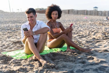 Sahilde kızgın genç bir çift, akıllı telefonlu bir kız arkadaş, erkek arkadaşının telefonuna bakan bir kız, erkek arkadaşının akıllı telefonunu gözetleyen ve gözetleyen genç bir kadın.