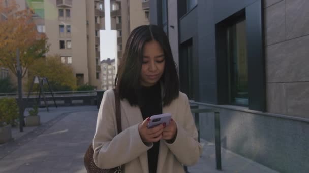 Fiatal felnőtt ázsiai nő gépel az okos telefonján, és félrenéz, miközben sétál a városban — Stock videók