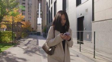 Genç Asyalı kadın şehirde yürürken akıllı telefonuna göz atıyor.