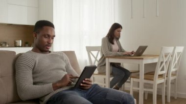 Karışık ırk çifti evde takılıyor, tablet üzerinde internette geziniyor ve evde bir dizüstü bilgisayar üzerinde çalışıyor.