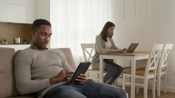 Gemengde race paar chillen thuis, surfen op het web op een tablet en werken aan een laptop thuis — Stockvideo