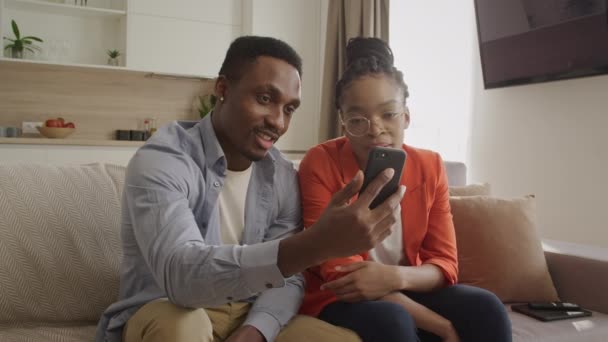 Jovem adulto casal afro-americano sentado em um sofá e acenando com seus amigos durante uma chamada de vídeo — Vídeo de Stock