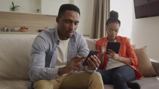 Dva mladí afroameričtí dospělí lidé se doma ochlazují a surfují na webu na telefonech a tabletách — Stock video