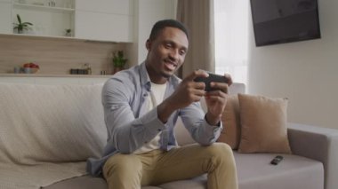 Mutlu gülümseyen Afrikalı Amerikalı adam dairesinde akıllı telefonuyla oynuyor.