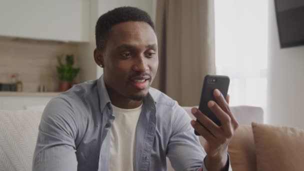 Feliz chico afroamericano en una videollamada y agitando su mano en su apartamento usando un teléfono inteligente — Vídeos de Stock